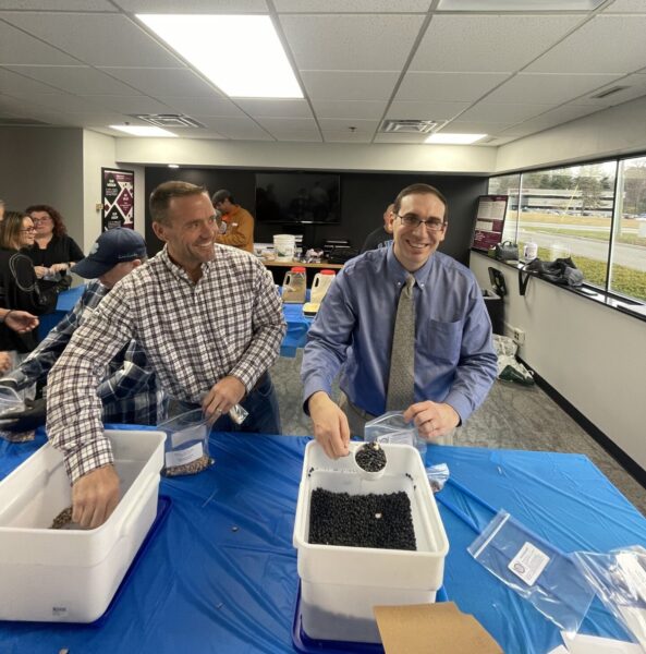 Two men in business casual attire, including Anthony Bordoley on the right, sorting small black items into containers on a blue-covered table in a bright office room.